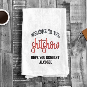 以下が含まれることがあります： 黒と赤の文字で「Welcome to the shitshow. Hope you brought alcohol.」と書かれた白いキッチンタオルです。