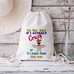 May include: A white drawstring bag with the embroidered text "I'm Not Sure If I ATTRACT Crazy or if I make them that way" and a cartoon chicken. A straw hat and a glass vase are in the background.