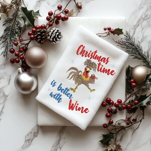 May include: White kitchen towel with red and green holly sprigs and berries. The towel features an embroidered design of a rooster holding a glass of wine with the text "Christmas time is better with Wine".