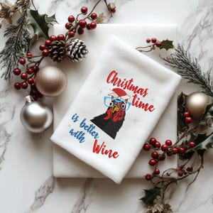 May include: White kitchen towel with red and blue embroidered text that reads "Christmas time is better with Wine". A cartoon chicken wearing glasses and a Santa hat is embroidered on the towel.
