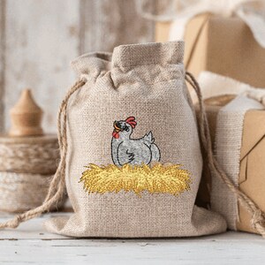 May include: A small burlap bag with a drawstring closure, featuring an embroidered design of a gray chicken sitting in a nest of yellow hay.