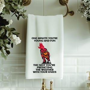 Può includere: Asciugamano bianco con il testo "ONE MINUTE YOU'RE YOUNG AND FUN" e "THE NEXT YOU'RE PREDICTING THE WEATHER WITH YOUR KNEES". L'asciugamano presenta una grafica di gallo colorata. L'asciugamano è appeso a un portasciugamani dorato.