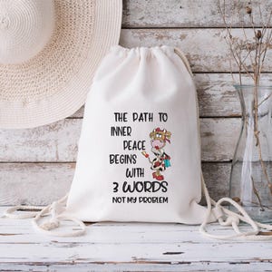 May include: White drawstring bag with a black and white cow cartoon illustration. The text on the bag reads "The path to inner peace begins with 3 words: Not my problem."