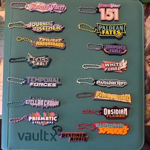 May include: A collection of sixteen colorful rubber keychains featuring various Pokémon trading card set logos, including Scarlet & Violet, 151, Journey Together, Paldean Fates, Twilight Masquerade, Shrouded Fable, Black Bolt, White Flare, Temporal Forces, Paradox Rift, Stellar Crown, Paldea Evolved, Prismatic Evolutions, Obsidian Flames, Surging Sparks, and Destined Rivals, each attached to a small metal ball chain.