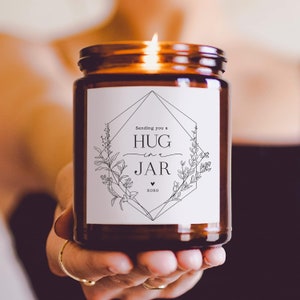 May include: A brown glass candle jar with a white label that says "Sending you a hug in a jar xoxo". The label has a geometric design with floral accents.