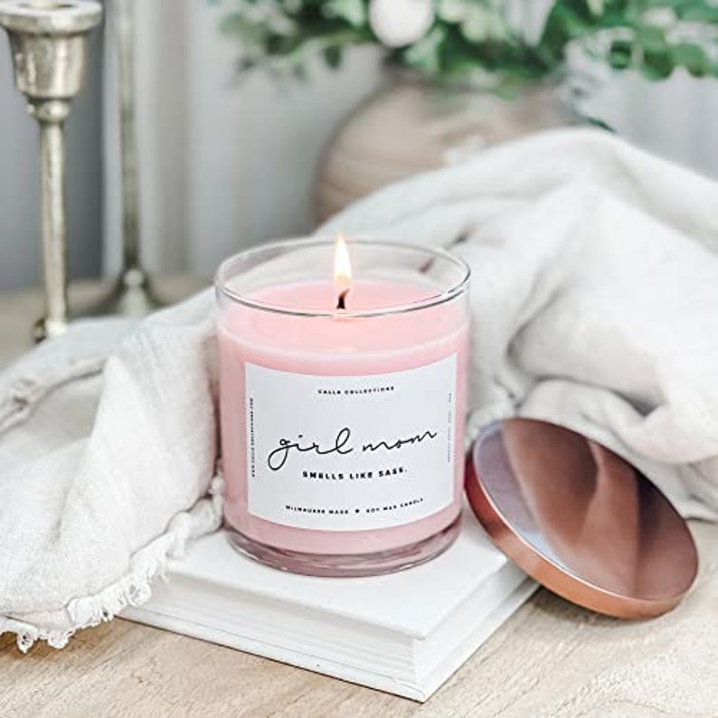 Pu&ograve; includere: Una candela di vetro rosa con un'etichetta bianca che recita "Girl Mom Smells Like Sass". La candela &egrave; accesa e ha un coperchio dorato.