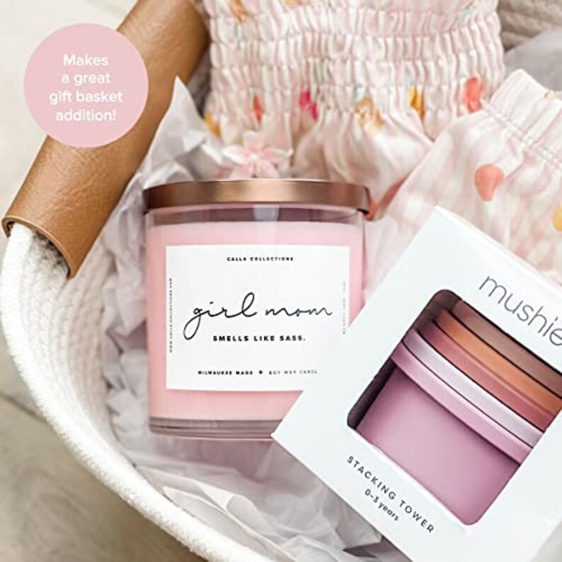 Pu&ograve; includere: Un cesto di vimini bianco pieno di una candela rosa con un coperchio dorato e un'etichetta bianca che recita "girl mom smells like sass." La candela &egrave; accanto a una scatola bianca con il testo "mushie stacking tower 0-3 years."