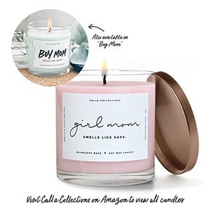 Pu&ograve; includere: Una candela di cera di soia rosa in un barattolo di vetro trasparente con un'etichetta bianca che recita "Calla Collections girl mom SMELLS LIKE SASS. MILWAUKEE MADE SOY WAX CANDLE". La candela &egrave; su una superficie bianca con un coperchio dorato.