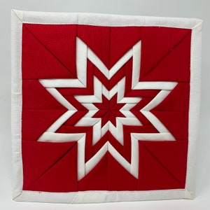 以下が含まれることがあります： 赤と白のキルティングされた星型の鍋つかみ。星は、生地の正方形をパターンに合わせて縫い合わせたものです。