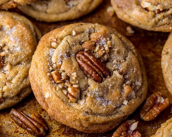 May include: Close-up of several golden-brown cookies, each topped with pecan halves and chopped pieces. The cookies have a slightly textured surface and are arranged on a rustic, golden-toned baking sheet. Some cookies have a dusting of sugar.