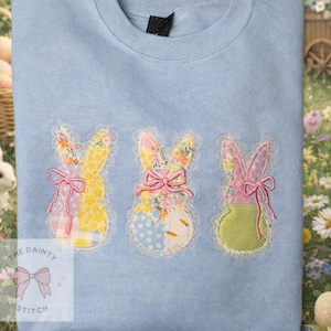 May include: Light blue sweatshirt featuring three appliqued bunny rabbits with floral and patchwork designs. Each bunny has a pink bow and is outlined with lace. The Dainty Stitch logo is in the lower left corner.