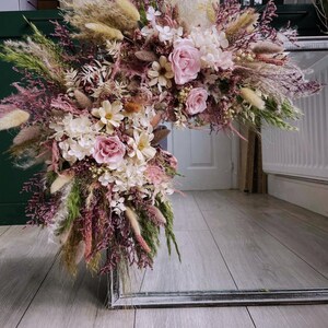 Peut inclure: Une grande composition florale composée de fleurs séchées et préservées dans des tons rose poudré, crème et vert doux. Le bouquet comprend des roses, des hortensias, de l'herbe de la pampa et diverses herbes plumeuses sur un cadre métallique.