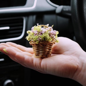 May include: A small woven basket brimming with dried flowers in green and purple hues. The basket is held in a hand, with a car interior in the background. The basket has a small handle and is made of a light brown material.