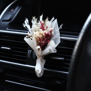 May include: A small bouquet of dried flowers in shades of white, cream, and burgundy, tied with a white ribbon, is displayed in a car's air vent. The flowers are arranged in a clear plastic bag and are secured to the vent with a clip.