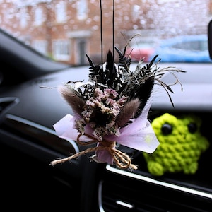 May include: A car air freshener with a bouquet of dried flowers in pink, black, and beige, wrapped in lavender paper and tied with twine. A green crocheted frog is visible in the background. The car window is visible.