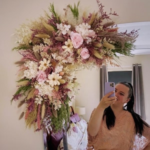 May include: A large, asymmetrical floral arrangement attached to the corner of a rectangular mirror. The arrangement features dried and preserved flowers in shades of dusty pink, cream, white, and sage green, including roses, hydrangeas, and grasses.