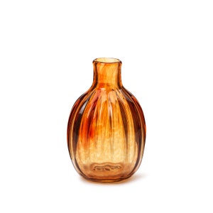 Puede incluir: Un jarrón pequeño de vidrio color ámbar con un diseño acanalado. El jarrón es redondo y tiene una abertura estrecha en la parte superior.