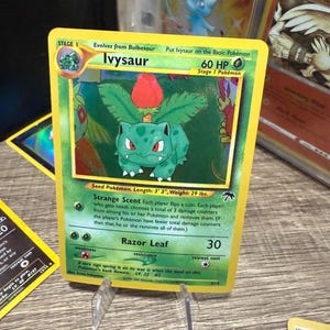 Könnte beinhalten: Eine Pokémon-Sammelkarte mit Ivysaur, einem grünen, pflanzenartigen Wesen mit einer roten Blume auf dem Rücken. Die Karte hat einen gelben Rand und enthält Text wie "Razor Leaf". Die Karte wird auf einem transparenten Ständer präsentiert.