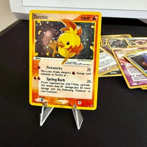 May include: A yellow and orange Pokemon trading card featuring Torchic, a fire-type Pokemon. The card has a 70HP rating and features two attacks: Fireworks and Spring Back. The card is set against a sparkly background.