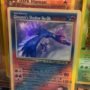 Può includere: Una carta Pokémon olografica con Giovanni's Shadow Ho-Oh, un Pokémon uccello blu e viola con un occhio rosso. La carta ha un valore di 200 PV e presenta il testo "Giovanni's Shadow Ho-Oh" e "200 PV".