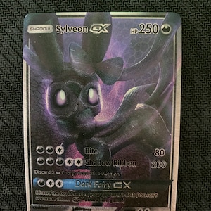 May include: A holographic Pokemon trading card featuring Sylveon GX, a purple and black fairy Pokemon with large eyes. The card has a HP of 250 and features three attacks: Bite, Shadow Ribbon, and Dark Fairy GX.