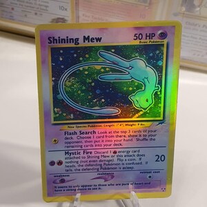 May include: A holographic Pokemon trading card featuring Shining Mew, a light blue and green mythical Pokemon with a star-filled background. The card text reads "Flash Search" and "Mystic Fire".