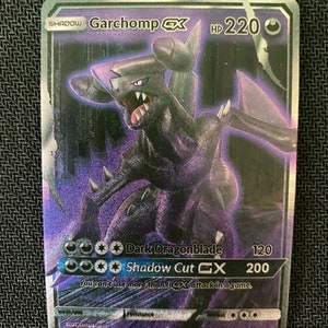 Puede incluir: Una carta de comercio de Pokemon holográfica con Garchomp GX, un Pokemon de tipo dragón de color morado oscuro y negro con ojos rojos. La carta tiene un fondo negro con un borde plateado y presenta el texto "SHADOW Garchomp GX HP 220". La carta tiene dos ataques: "Dark Dragonblade" por 120 de daño y "Shadow Cut GX" por 200 de daño. La carta también tiene una regla que establece "No puedes usar más de 1 ataque GX en un juego."