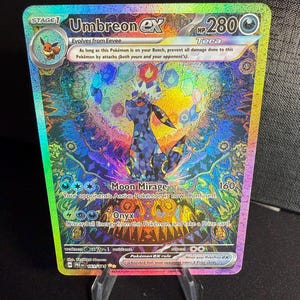 以下が含まれることがあります： ホログラフィックなポケモンカードゲームのカードで、ウmbreon EXが描かれています。ウmbreon EXは、暗い青と紫色のポケモンで、黄色い目をしています。カードにはテラマークと「このポケモンがベンチにいる限り、このポケモンへの攻撃によるダメージをすべて防ぐ（あなたの攻撃と相手の攻撃の両方）。 」というテキストが書かれています。カードには、ムーンミラージュとオニキスという2つの技も書かれています。