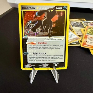 Può includere: Una carta Pokémon Umbreon del Gioco di Carte Collezionabili Pokémon. La carta presenta un Umbreon nero e blu con occhi gialli. La carta ha un Poké-Potere chiamato Raggio Oscuro e un attacco chiamato Attacco Finto. La carta è del set 17/17.