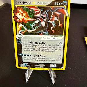 May include: Charizard Pokemon card from the Delta Species set. The card is yellow with a holographic image of Charizard. The card has a 90HP rating and the text "Rotating Claws" and "Dark Swirl" are visible.