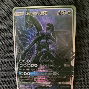 May include: A holographic Pokemon trading card featuring Sceptile GX, a dark green and purple creature with glowing eyes. The card has a black background and features the text "Shadow Cut 80" and "Black Poison 300".
