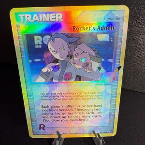 May include: A holographic Pokemon trading card featuring Rocket's Admin. The card is a Trainer card and features a blue and white illustration of a character with a purple cat. The card text reads "You can play only one Supporter card each turn. When you play this card, put it next to your Active Pokemon. When your turn ends, discard this card. Each player shuffles his or her hand into his or her deck. Then, each player counts his or her Prize cards left and draws up to that many cards. (You draw your cards first.)"