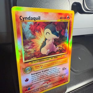 Puede incluir: Una tarjeta Pokémon holográfica de Cyndaquil. La tarjeta presenta una imagen de dibujos animados de Cyndaquil, un pequeño Pokémon bípedo de tipo fuego. La tarjeta tiene un borde amarillo e incluye texto como "Emergency Eruption" y "40 HP".