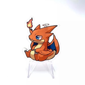 Può includere: Un piccolo drago di cartone animato arancione e blu con una fiamma sulla coda. Il drago è seduto su un supporto in acrilico trasparente.