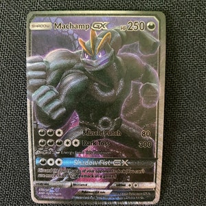 May include: A holographic Pokemon trading card featuring Machamp GX, a fighting type Pokemon with a dark purple and black color scheme. The card has a black background with a silver border and features the text "SHADOW Machamp GX HP 250". The card also features three attacks: Muscle Punch, Dark Toss, and Shadow Fist GX.