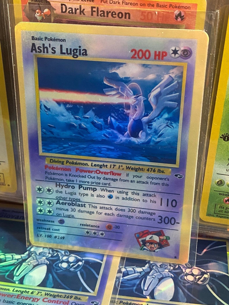 May include: A Pokemon card featuring Ash's Lugia. The card is blue and purple with a holographic finish. The card has a picture of Lugia in the background. The card has the following text: 'Ash's Lugia', '200 HP', 'Diving Pokemon. Lenght 17' 1', 'Weight: 476 lbs.', 'Pokemon Power: Overflow', 'Hydro Pump', 'Aeroblast', 'Weakness', 'Resistance', 'Retreat Cost', 'LV. 100 #249'