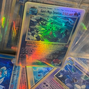 May include: A holographic Pokemon card featuring Gary's Mega Tyranitar with 250 HP. The card is green, blue, and purple with a holographic effect. The card text reads "Armor Pokemon. Length 6' 07", Weight: 562.2 lbs."