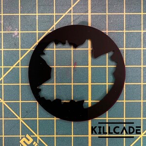 Puede incluir: Plantilla circular negra con un contorno irregular y dentado de una forma que se asemeja a un mapa. La plantilla est&aacute; sobre una superficie cuadriculada verde y amarilla.