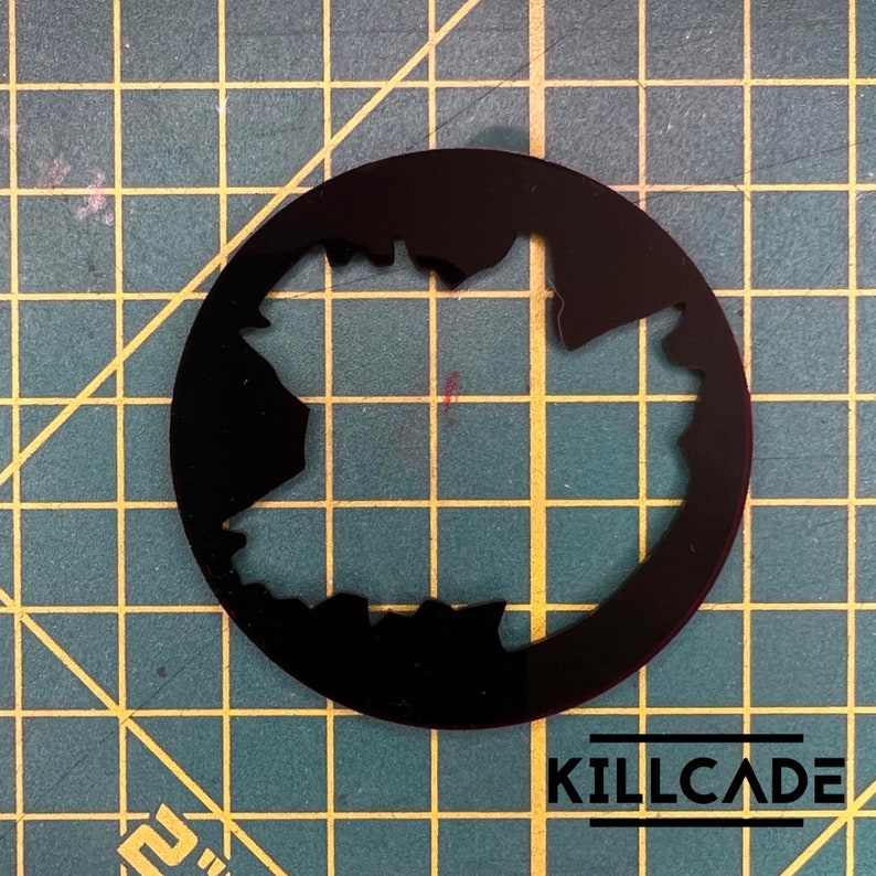 Puede incluir: Plantilla circular negra con un contorno irregular y dentado de una forma que se asemeja a un mapa. La plantilla est&aacute; sobre una superficie cuadriculada verde y amarilla.