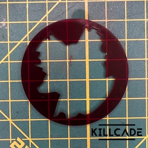 Puede incluir: Una plantilla circular de color rojo oscuro con una forma irregular y dentada recortada en el centro. La plantilla est&aacute; sobre un fondo de cuadr&iacute;cula verde.