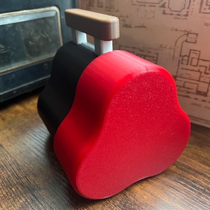 Könnte beinhalten: Eine rot-schwarze, 3D-gedruckte herzförmige Box mit einem Holzgriff oben. Die Box steht auf einer Holzoberfläche.