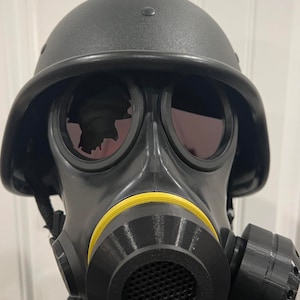 Puede incluir: Una m&aacute;scara de gas negra con una banda amarilla alrededor de la boquilla. La m&aacute;scara tiene dos grandes piezas de ojos redondas y un casco unido a la parte superior.