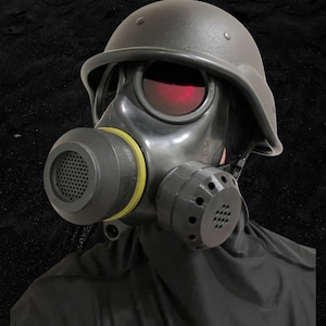 Puede incluir: Una m&aacute;scara de gas negra con un casco negro y un sello amarillo alrededor de la boquilla. La m&aacute;scara tiene una visera tintada de rojo y un filtro negro en el lateral.