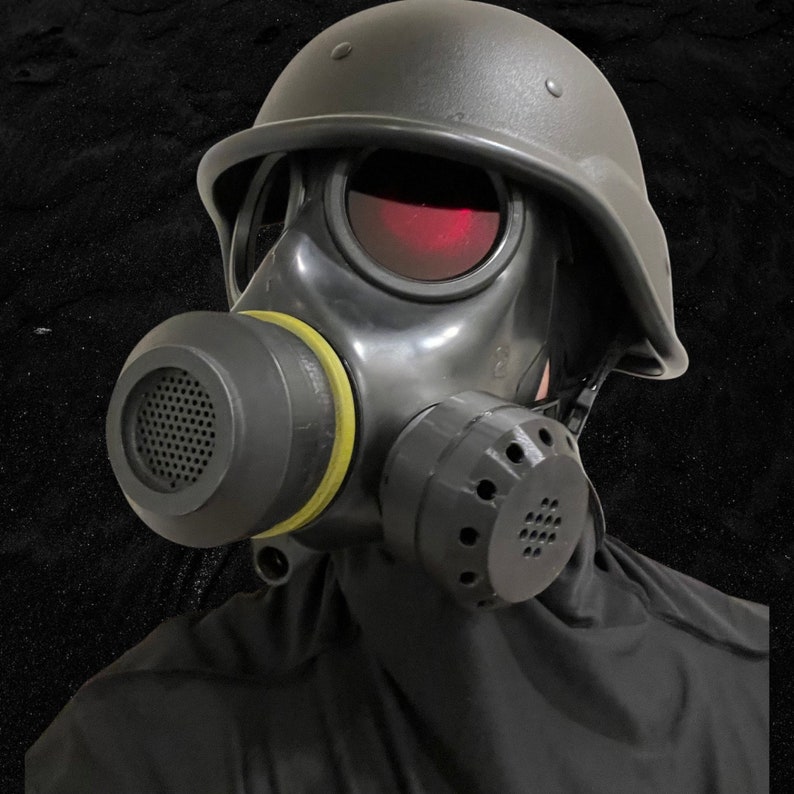 Puede incluir: Una m&aacute;scara de gas negra con un casco negro y un sello amarillo alrededor de la boquilla. La m&aacute;scara tiene una visera tintada de rojo y un filtro negro en el lateral.