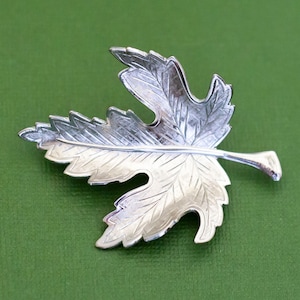 Puede incluir: Broche plateado en forma de hoja. El broche tiene vetas detalladas y una superficie texturizada, con una combinación de colores plateados claros y oscuros. El broche está sobre un fondo verde.