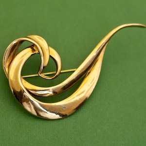 Puede incluir: Broche dorado con un diseño abstracto y ondulado. La broche presenta una forma curva y fluida con un alfiler en la parte posterior para sujetar. El diseño tiene un acabado pulido y está sobre un fondo verde.