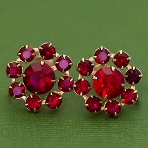 Pode incluir: Um par de brincos dourados com detalhes em strass vermelho. Os brincos têm a forma de uma flor com vários strass vermelhos.
