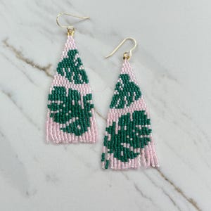 May include: A pair of beaded earrings with a pink background and a green leaf pattern. The earrings have gold-colored hooks and a triangular shape. The earrings are on a white marble surface.