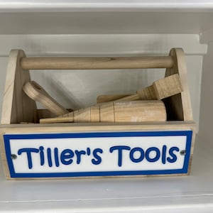 May include: A wooden toy toolbox with a blue and white sign that says "Tiller's Tools". The toolbox is filled with wooden tools, including a hammer, a saw, and a screwdriver.