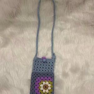 May include: A light blue crochet crossbody bag with a long strap. The bag features a floral design in shades of purple, green, and cream. A button closure secures the top flap. The bag is displayed on a white, fluffy surface.